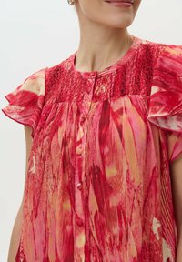 Blumenkleid in Rot- und Rosatönen, mit kurzen Rüschensäumen, Knopfleiste und texturiertem Oberteil mit Smocking-Details. Leichtes Material.