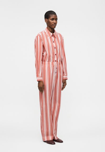 Γυναίκα με ταιριαστό κόκκινο και λευκό πουκάμισο με κάθετες ρίγες και φαρδιά παντελόνια με σκούρα παπούτσια, στέκεται μπροστά σε απλό φόντο.