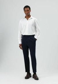 Chemise blanche boutonnée à manches longues, associée à un pantalon bleu marine. Le pantalon présente une coupe cintrée et des poches latérales. Des mocassins marron complètent le look.