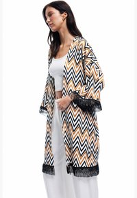 Kvinde iført en kimono med et flerfarvet zigzag-mønster og sort frynse over en hvid tanktop og brede hvide bukser, stående i profil.
