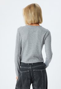 Graues Oberteil mit langen Ärmeln und Rundhalsausschnitt, aus weichem Stoff. Getragen mit hochgeschnittenen, dunkelgrauen, weiten Hosen, die große Gesäßtaschen haben.