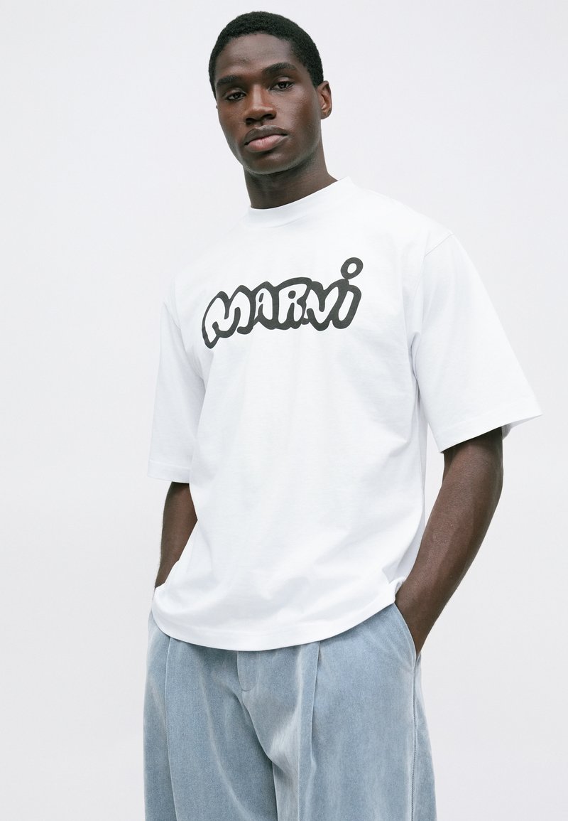 Homme portant un T-shirt oversized blanc avec le logo "Marni" en noir, un pantalon ample bleu clair, se tenant avec les mains dans les poches sur fond blanc uni.