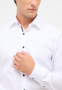 Hvid skjorte med knaplukning i et struktureret stof, sorte knapper og kontrasterende sort kant langs kraven. Langærmede med manchet detaljer.