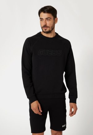 Črna kapuca z raglan rokavi, ki se ponaša z teksturiranim logotipom "GUESS" čez prsni del. Skupaj z črnimi kratkimi hlačami, na katerih je prikazan isti logotip.