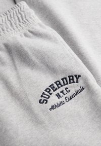 Zbliżenie na szare spodnie dresowe z elastycznym paskiem i czarnym haftowanym napisem "SUPERDRY N.Y.C Athletic Essentials" na obszarze kieszeni.