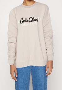 Beige Sweatshirt mit geripptem Halsausschnitt und Bündchen, mit schwarzem gesticktem "Carlo Colucci Milano 1978" Logo auf der Vorderseite. Jeans aus Denim sichtbar.