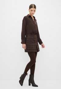 Blusa button-up marrone abbinata a una gonna a quadri marrone, collant scuri e stivaletti. Tessuto liscio, design strutturato e motivi sottili.