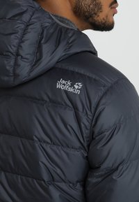 Homme portant une veste matelassée noire Jack Wolfskin avec capuche, vue de dos et de profil.
