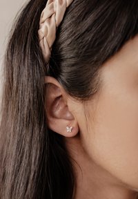 Boucles d'oreilles puces en argent en forme de papillon avec de petites pierres claires, posées sur un teint de peau clair, cheveux coiffés avec un bandeau en satin tressé.
