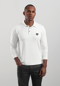 Langärmliges weißes Polohemd mit geknöpftem Kragen und schwarzem Emblem auf der Brust. Glatter Stoff mit figurbetontem Schnitt und minimalistischem Look.