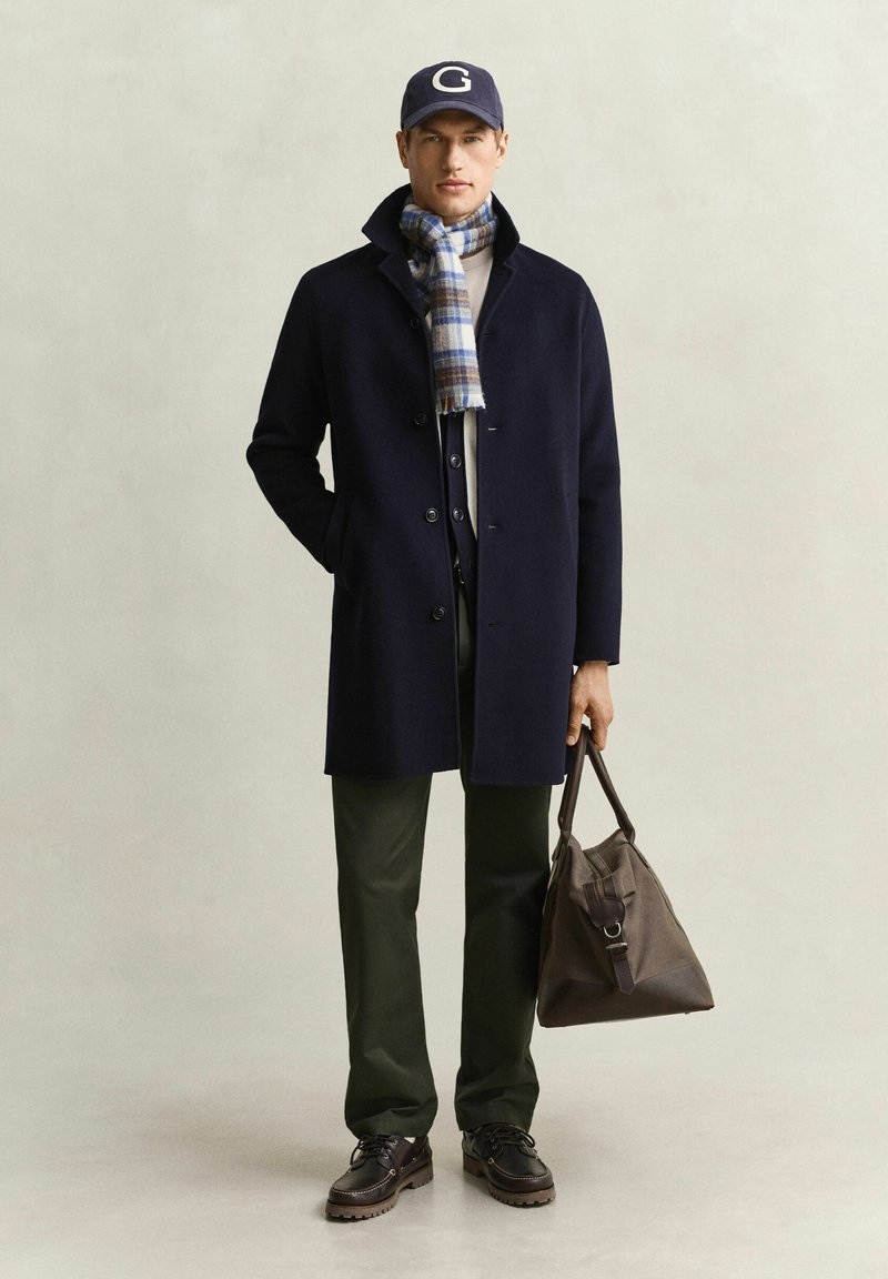 Homme portant un manteau bleu marine, une écharpe à carreaux, une casquette bleu marine, un pantalon foncé et des chaussures marron, tenant un sac en cuir marron dans la main droite sur un fond uni.
