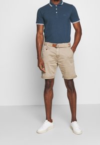 Námořnická polo košile s bílými detaily v kombinaci s béžovými šortkami a tkaným opaskem. Bílí tenisky dotváří celý outfit. Jednoduchý a neformální design.