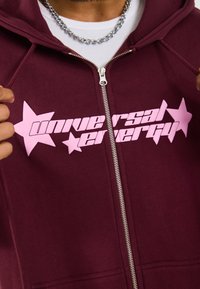 Haut à zip bordeaux, arborant des graphiques d'étoiles roses et le texte "Énergie Universelle". Tissu doux avec une texture lisse et fermeture éclair en métal.