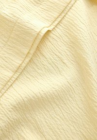 Helles gelbes Gewebe mit einer strukturierten, zerknitterten Oberfläche, das ein dezentes Gittermuster aufweist und sanfte Falten bildet.