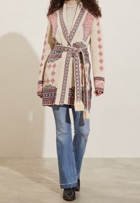 Mujer con un cardigan beige con patrón y cinturón sobre un cuello alto color crema, combinado con jeans azul claro y botas negras hasta el tobillo.