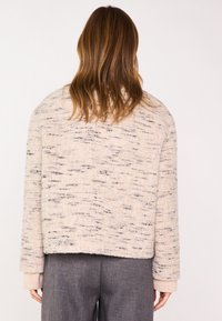 Veste tricotée beige avec un motif texturé et une coupe décontractée. Manches longues avec des poignets côtelés. Portée sur un pantalon gris.