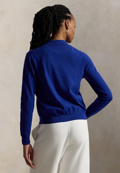 Μπλε πλεκτό πουλόβερ με κλασικό γιακά, μακριά μανίκια και ριμπ τελείωμα· λεία υφή, εφαρμοστή σχεδίαση και ελάχιστες ραφές.