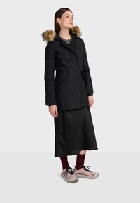 Čierna parka bunda s kapucňou podšitú kožušinou, skombinovaná s čiernou saténovou sukňou a ružovými teniskami. Má upravený strih a dve predné vrecká.