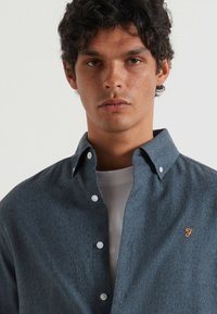 Męska koszula z guzikiem w ciemnoniebieskim kolorze, z teksturowanym wykończeniem, z białymi guzikami i pomarańczowym logo na lewej piersi.