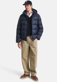 Marineblå pufferjakke med hætte, lynlås foran og sidelommer; båret over en marineblå sweater og en ternet skjorte. Beige bukser og brune sko.
