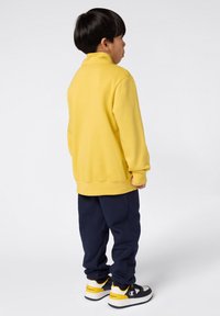 Sudadera de polar amarilla, con cuello alto y mangas largas. Combinada con pantalones de chándal azul marino y zapatillas negras/blancas con detalles amarillos.