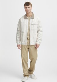 Witte canvas jas met een bruine kraag, voorzien van een voorzak, gedragen met beige broek en witte sneakers. Model staat in het midden.