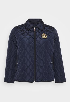 Veste matelassée marine avec col, fermeture éclair et un emblème décoratif sur la poitrine. Deux poches latérales sont visibles.