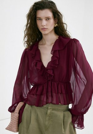 Žena s kučeravými vlasmi, oblečená v priehľadnej bordovej blúzke s volánmi, olivové zelené nohavice, zlaté kruhové náušnice, drží pletenú kabelku.