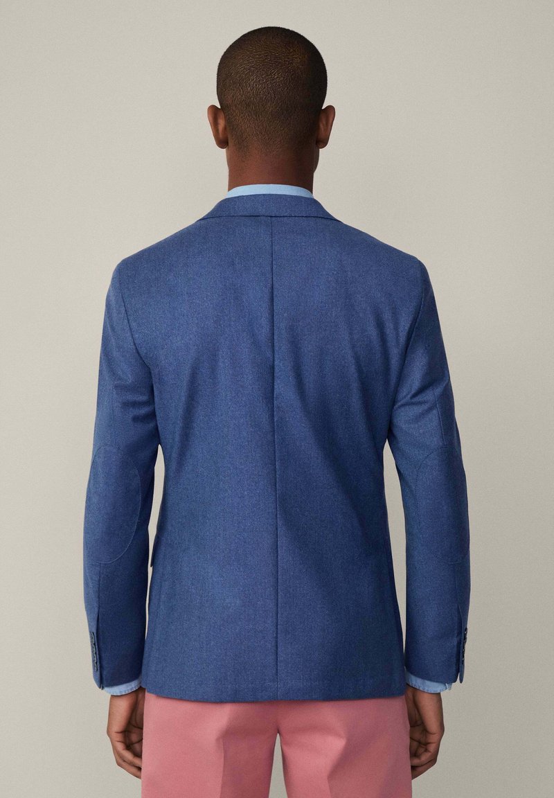 Donkerblauwe blazer met een gestructureerd ontwerp, met een enkele split, elleboogpatches en knopen aan de mouwen, gedragen over een lichtblauw overhemd.