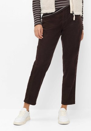 Person trägt dunkelbraune Hose mit geradem Bein, weiße Sneakers, gestreiftes Langarmshirt und cremefarbene Fleeceweste und steht vor weißem Hintergrund.
