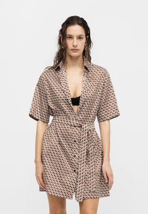 Kobieta z mokrymi włosami nosi krótki rękaw, sukienkę w geometryczny wzór z paskiem, na czarnym topie, stojąc na jednolitym białym tle.