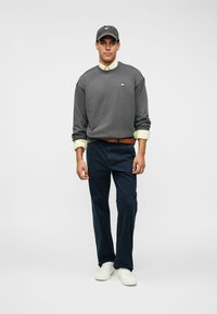 Sweatshirt cinza com punhos canelados, sobre uma camisa amarelo claro. Calças azul-escuro e ténis brancos, combinados com um boné cinza correspondente.