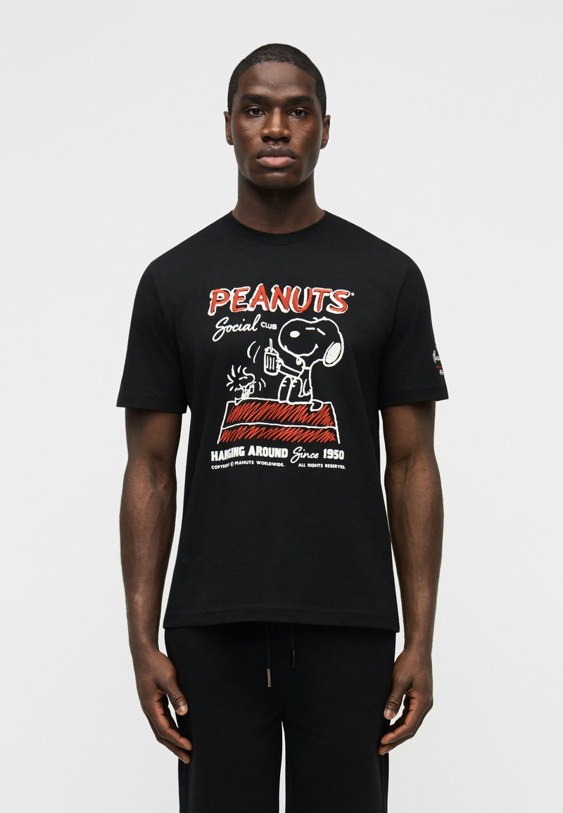 Άντρας που φοράει μαύρο μπλουζάκι Peanuts με τον Snoopy και τον Woodstock να κάθονται σε ένα κόκκινο σπιτάκι σκύλου με το κείμενο «Hanging Around Since 1950».