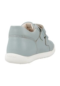 Chaussure en cuir gris avec une texture lisse, dotée de deux sangles réglables et d'une semelle en caoutchouc blanche. Des détails de couture subtils sont présents.
