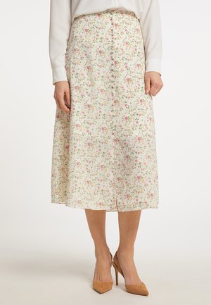 Frau trägt einen weißen Midirock mit kleinem rosa und gelbem Blumenmuster und beigen High Heels, steht vor einem schlichten Hintergrund.
