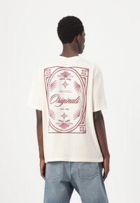 Kreemivärvi T-särk, mis on valmistatud puuvillast, seljal on suur punane lilleline graafika, millel on tekst "Originals" ja "EST. 1990". Lõdva lõikega.