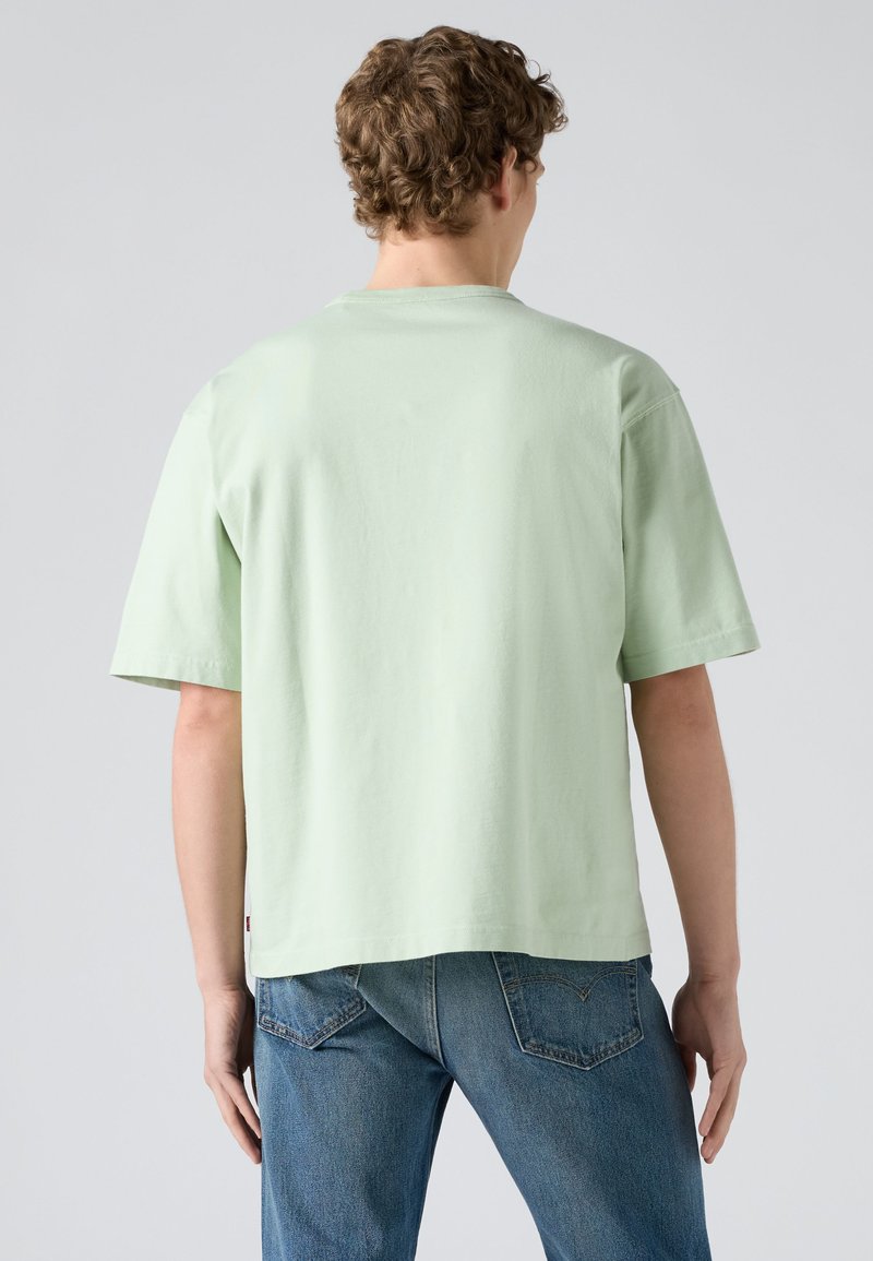 Jeune homme aux cheveux bouclés portant un T-shirt vert clair à manches courtes et un jean bleu, debout de dos sur un fond gris uni.