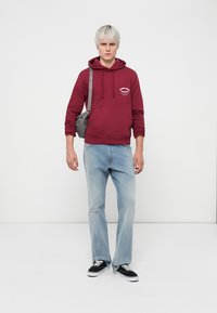 Haut à capuche bordeaux avec poche avant et logo, associé à un jean bleu clair et des baskets noires. Les accessoires comprennent un sac bandoulière gris.
