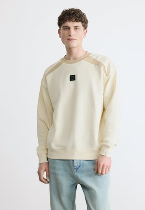 Tânăr cu păr creț, purtând un hanorac crem cu un mic patch negru cu logo, asortat cu blugi albastru deschis, pe un fundal alb.