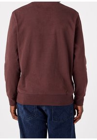 Uomo che indossa una felpa a maniche lunghe di colore marrone scuro e jeans in denim blu, visto da dietro.