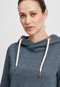 Mørkeblå hettegenser med høy hals, kremfargede snor, og brune lærdetaljer ved halsen. Glatt tekstur, avslappet design.