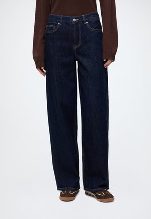 Persoon die donkerblauwe jeans met wijde pijpen, een bruine trui met lange mouwen en bruine instapschoenen met witte stiksels draagt, tegen een witte achtergrond.
