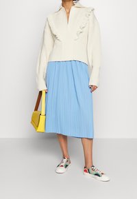 Maglione lavorato a maglia crema con colletto arricciato, gonna plissettata azzurra chiara e sneakers multicolori. Borsa gialla con dettagli marroni.