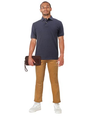 Man staat en draagt een navy poloshirt, mosterdkleurige broek, witte sneakers, en houdt een bruine leren polstas in zijn rechterhand tegen een witte achtergrond.