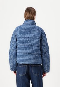 Vadderad blå jacka i denim med ett mönster av upprepande text. Hög krage, långa ärmar och en lätt kortare design.