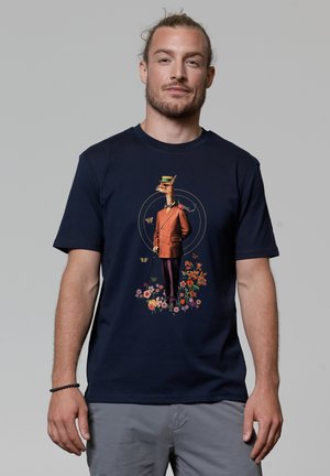 Marineblauw katoenen T-shirt met een speelse giraffe in een brown suit met bloemenaccenten, staande tussen kleurrijke bloemen en vlinders.