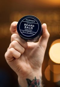 Mørkeblå cylindrisk blikdåse med Murdock Beard Balm, mærket med hvid tekst. Hånd der holder dåsen, med en synlig tatovering på håndleddet.