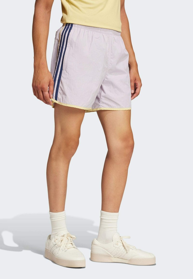 Lila Sportshorts mit gelbem Saum, die auf jeder Seite zwei marineblaue Streifen haben. Getragen mit weißen Turnschuhen und gerippten Socken.