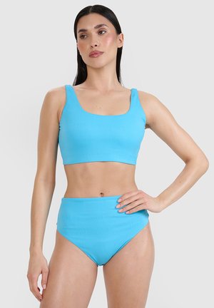 Frau mit langen dunklen Haaren, die einen hellblauen strukturierten zweiteiligen Badeanzug mit Crop-Top und hoch taillierten Hosen vor einfachem Hintergrund trägt.