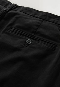 Black fabric pants feature a rear pocket with a stitched outline and a black button closure. The texture is smooth with a slight sheen.